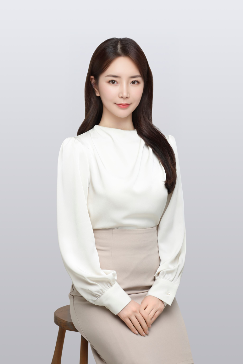 흰색 블라우스와 베이지색 치마를 입은 여성이 의자에 앉아 정면을 보고 있는 상반신 사진