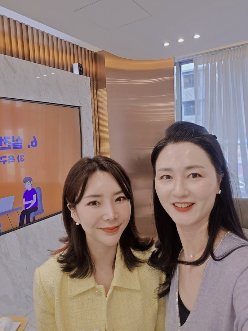 두 명의 여성이 실내에서 카메라를 보고 웃으며 찍은 셀카. 한 명은 노란색 재킷을 입고 있고 다른 한 명은 회색 상의를 입고 있다.